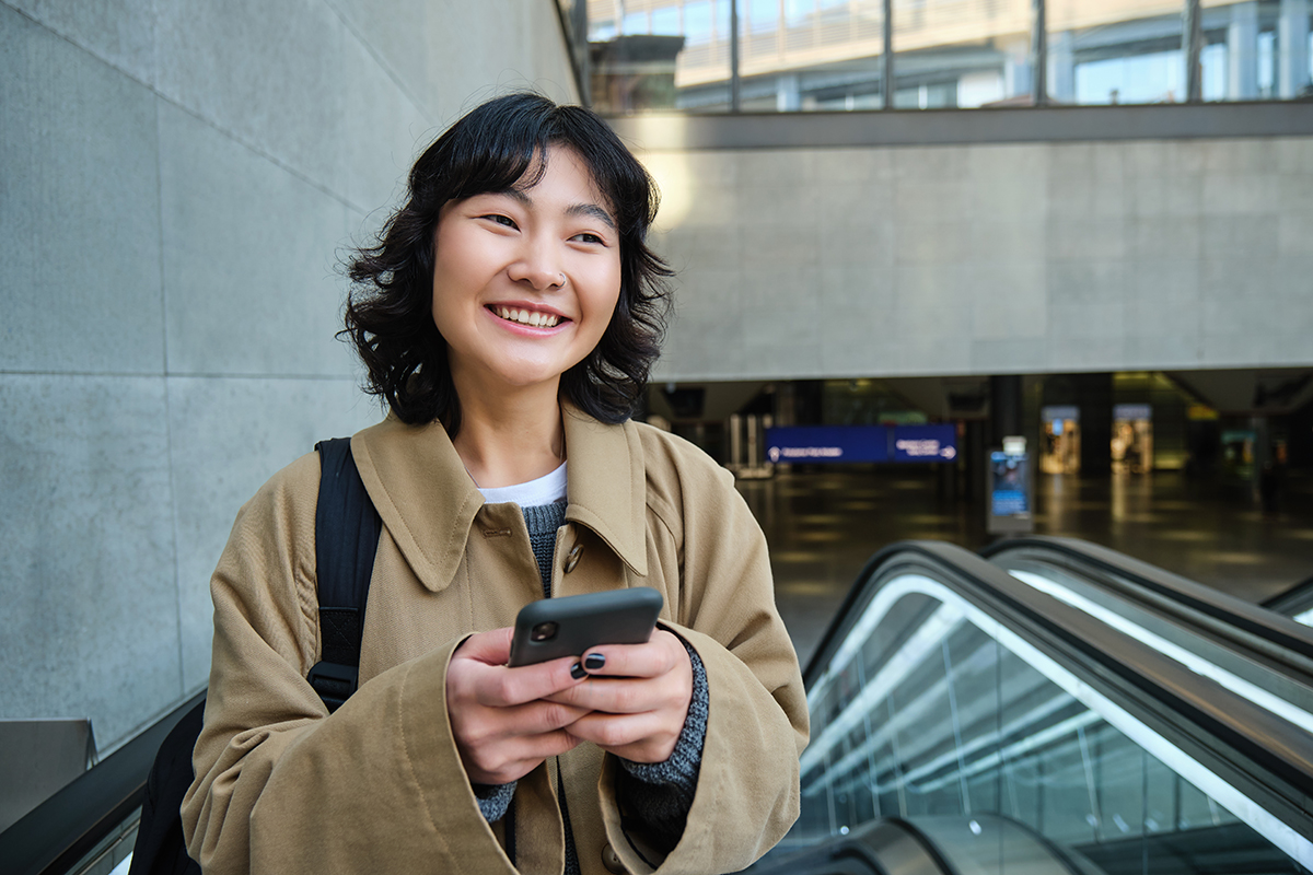Portrait of cute korean girl in trench coat, going up escalator, holds mobile phone and smiles pleased, commutes